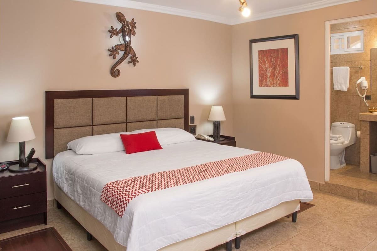 Habitación de hotel con una cama grande, una mesita de noche con lámpara, una mesita junto a la cama con teléfono, una obra de arte colgada en la pared y un baño visible a través de una puerta abierta.