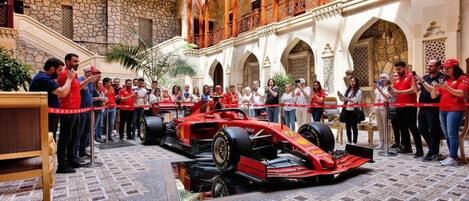 Une voiture de Formule 1 rouge se trouve dans une cour bordée de murs et d’arcades en pierre, et elle est entourée de personnes.