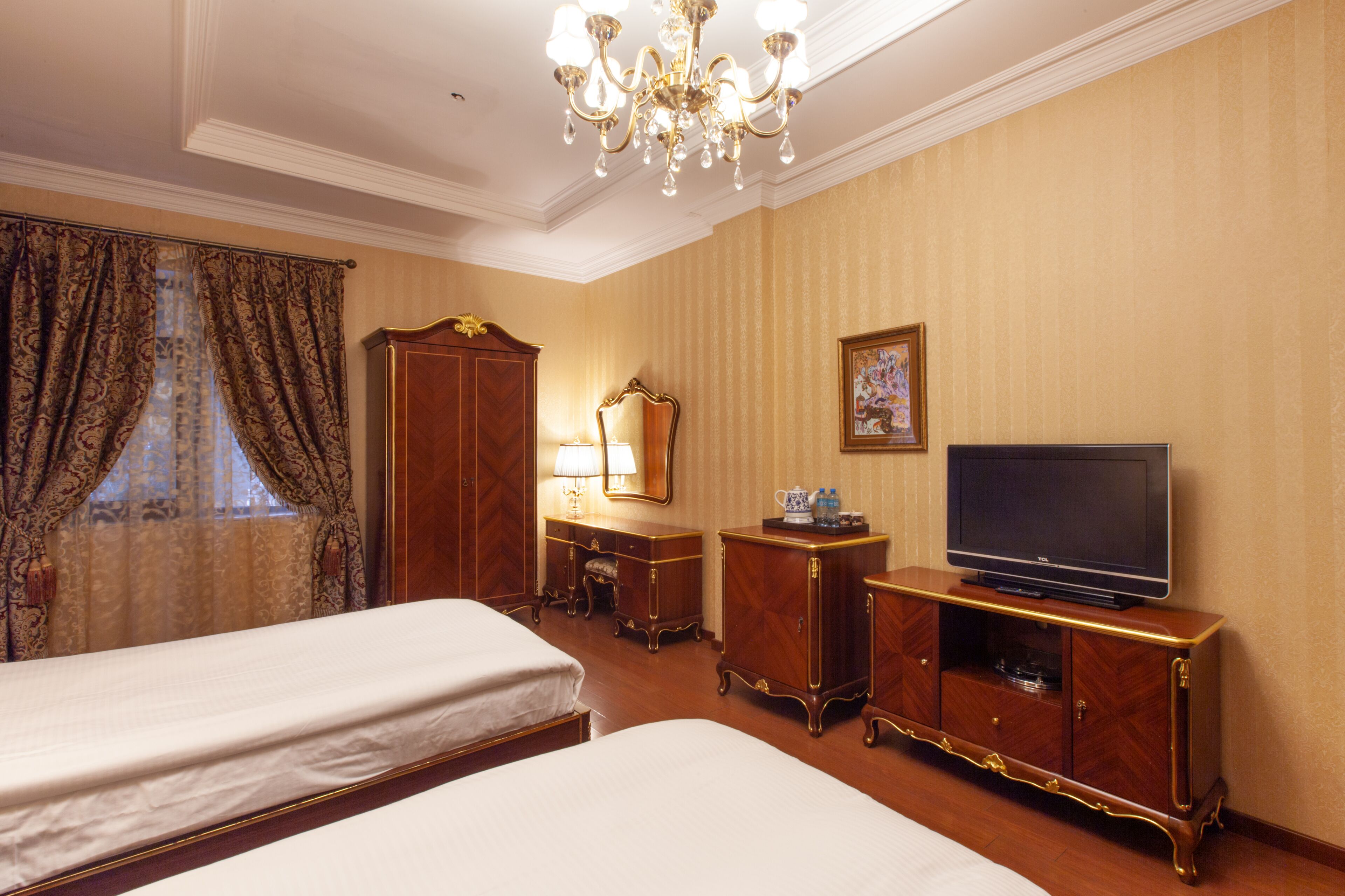 A hotel room with two beds, a television, a dresser, a mirror, and a chandelier.