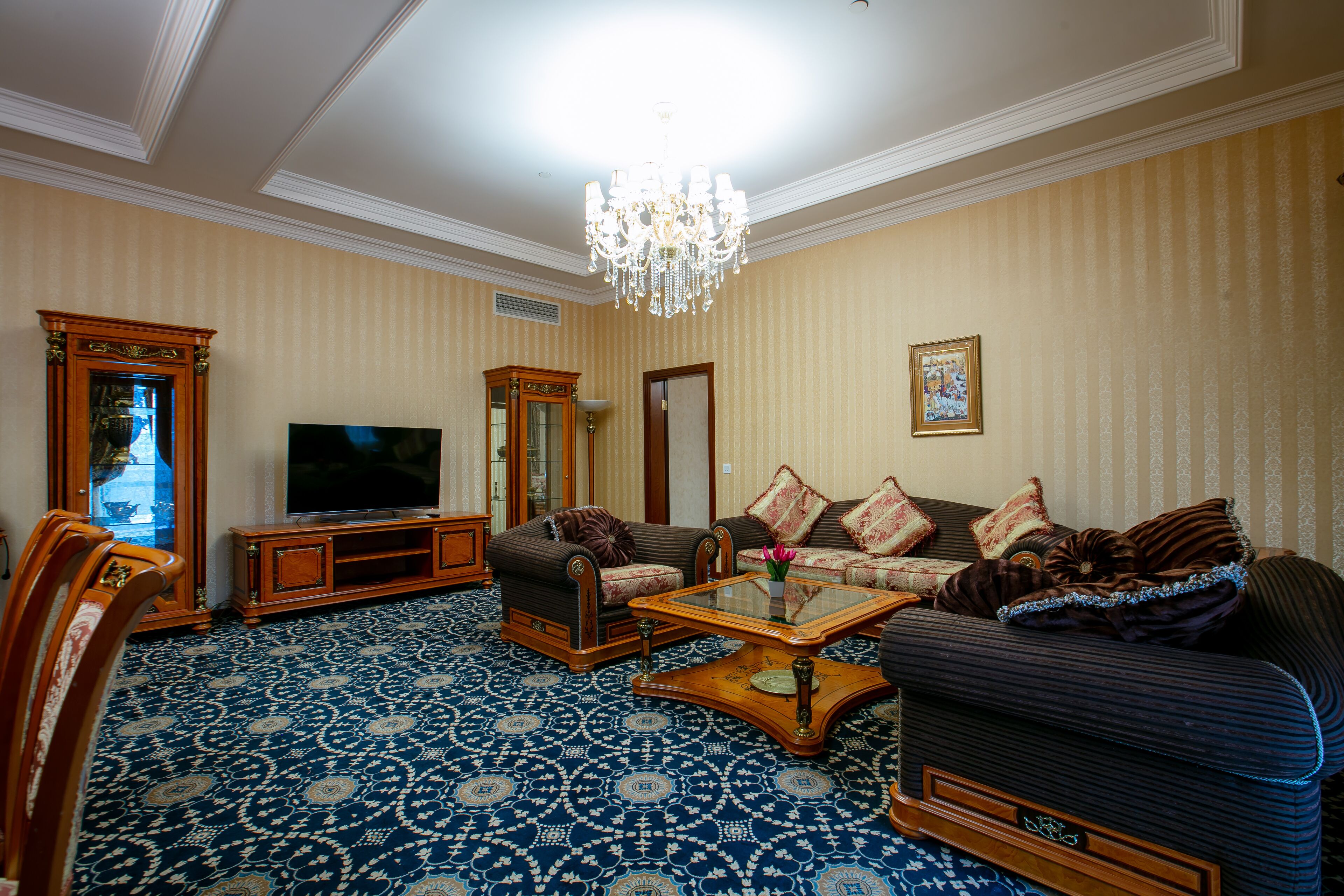 A spacious living room with a large television, a chandelier, and patterned carpet.