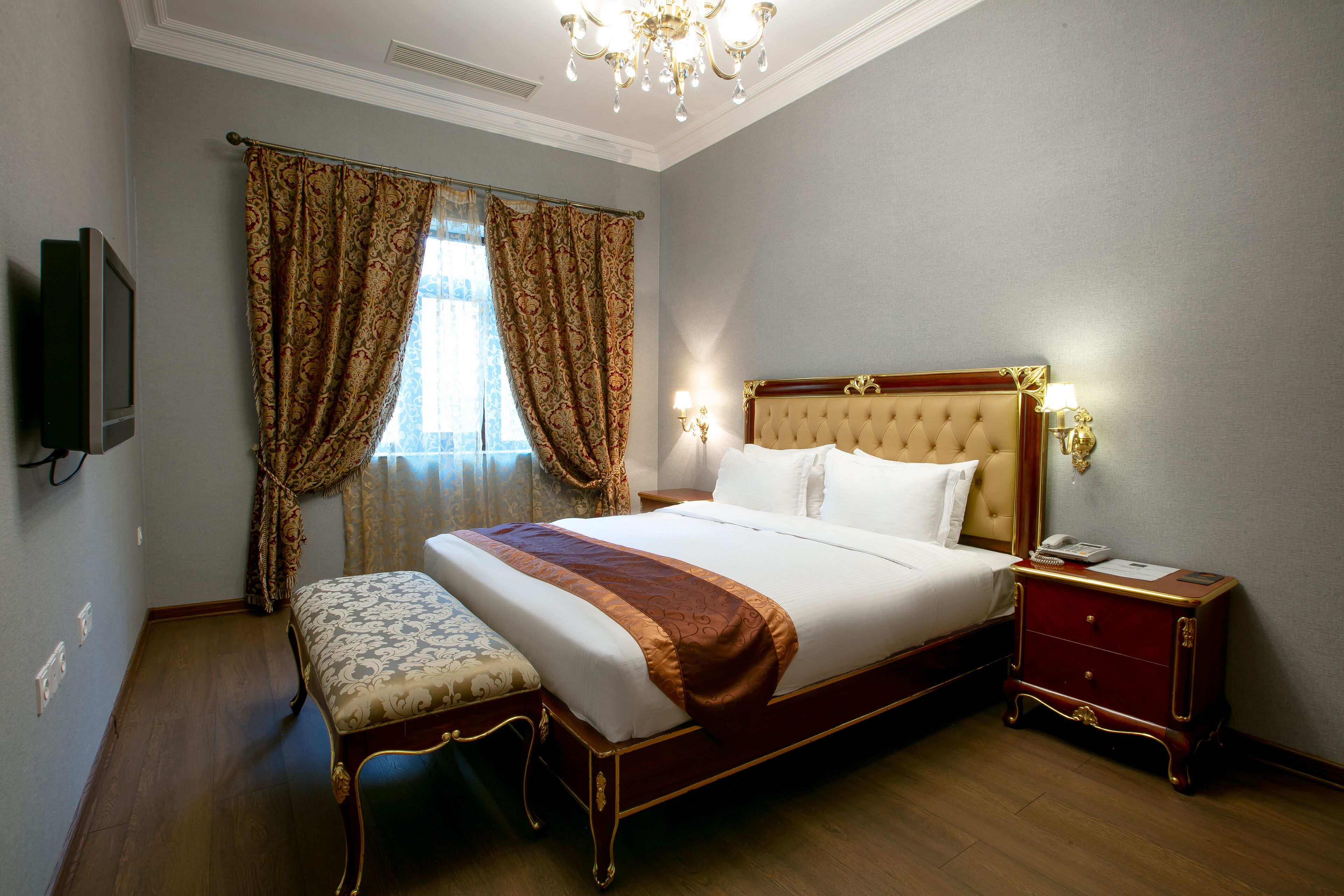 A hotel room with a large bed, a bedside table, a bench, and a television on the wall.