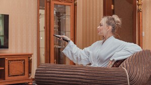 A person in a white robe holding a remote control in a room with a patterned carpet, a wooden cabinet, and a television. - Shah Palace Luxury Museum Hotel (Baku)