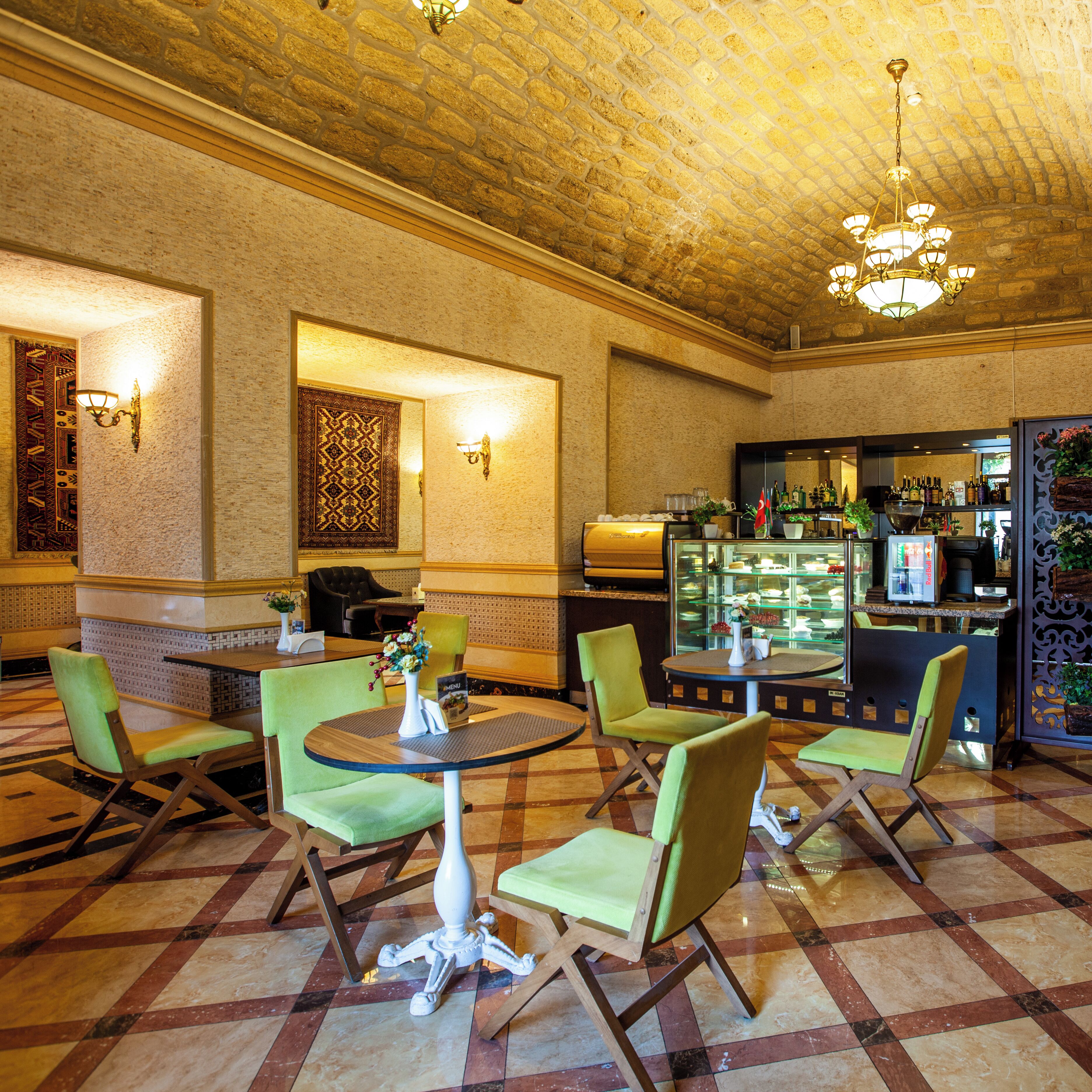 A hotel lobby with a checkered floor, green upholstered chairs, a round table with a vase of flowers, a bar area with shelves of bottles, and a patterned wall.