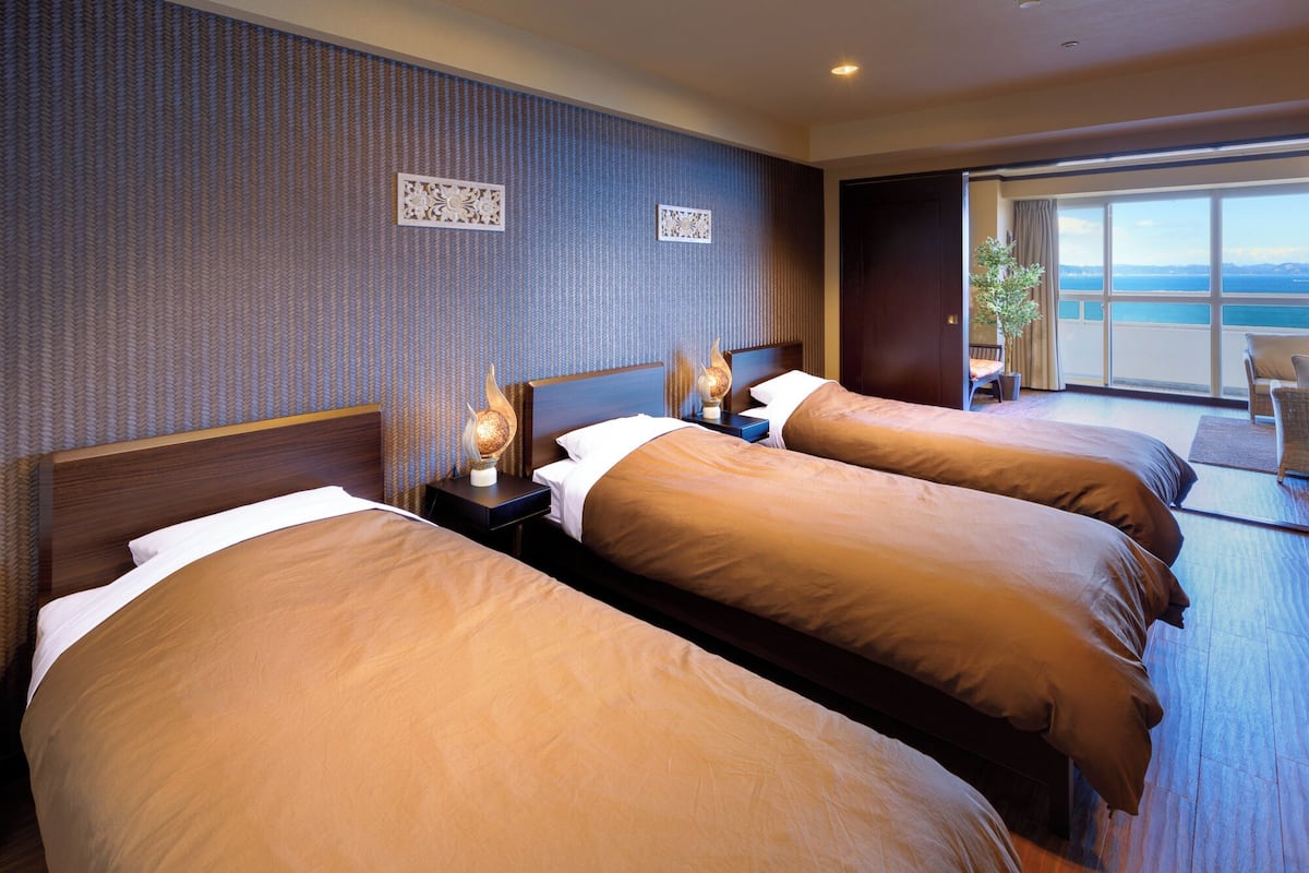 Een hotelkamer met twee bedden, een nachtkastje met een lamp en uitzicht op zee door grote ramen.