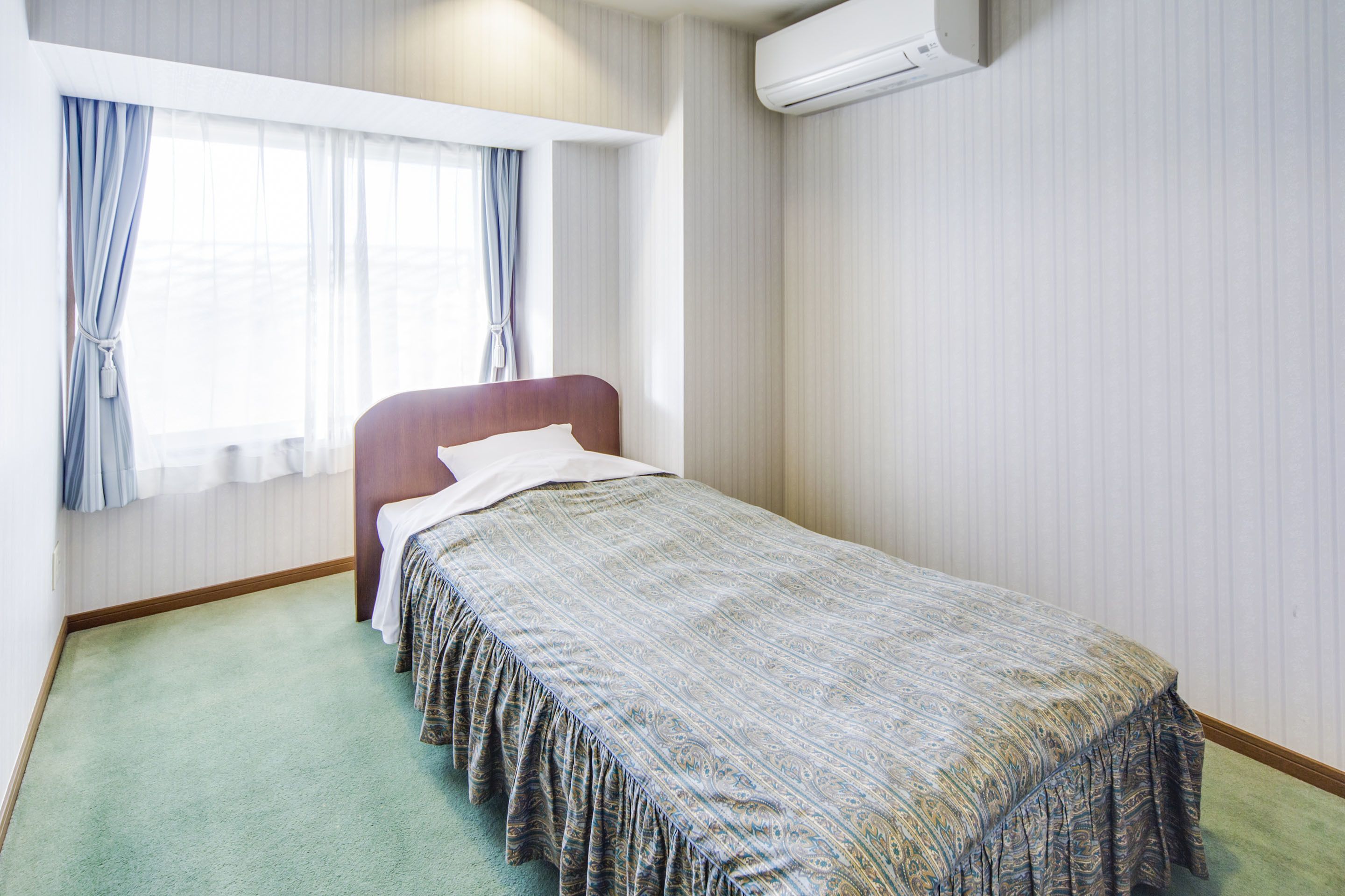 A single bed with a patterned bedspread, a window with curtains, and an air conditioning unit on the wall.