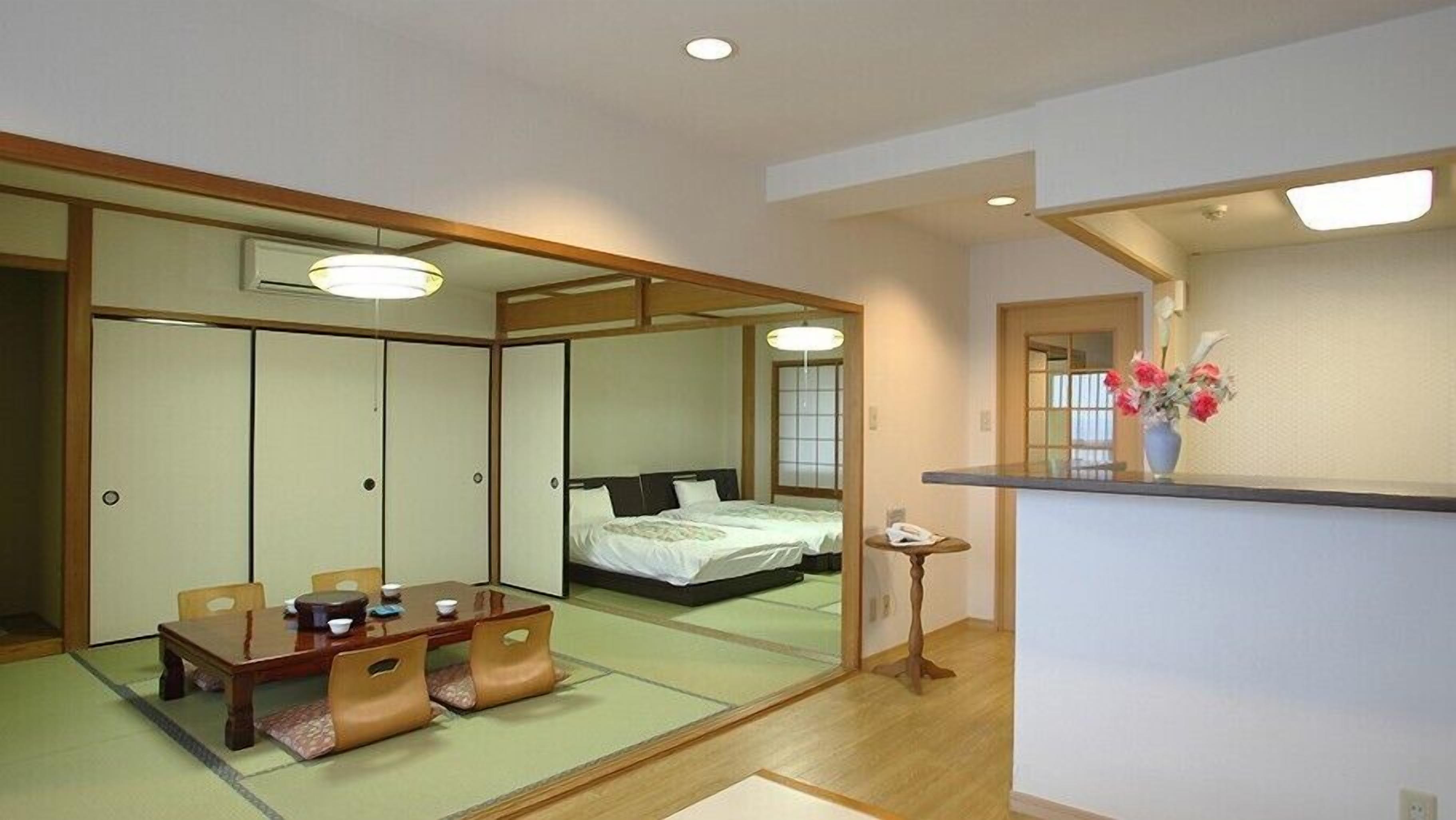 Een traditionele Japanse kamer met tatami vloeren, een lage tafel met kussens, een bed met een donkere hoofdeinde en schuifdeuren.
