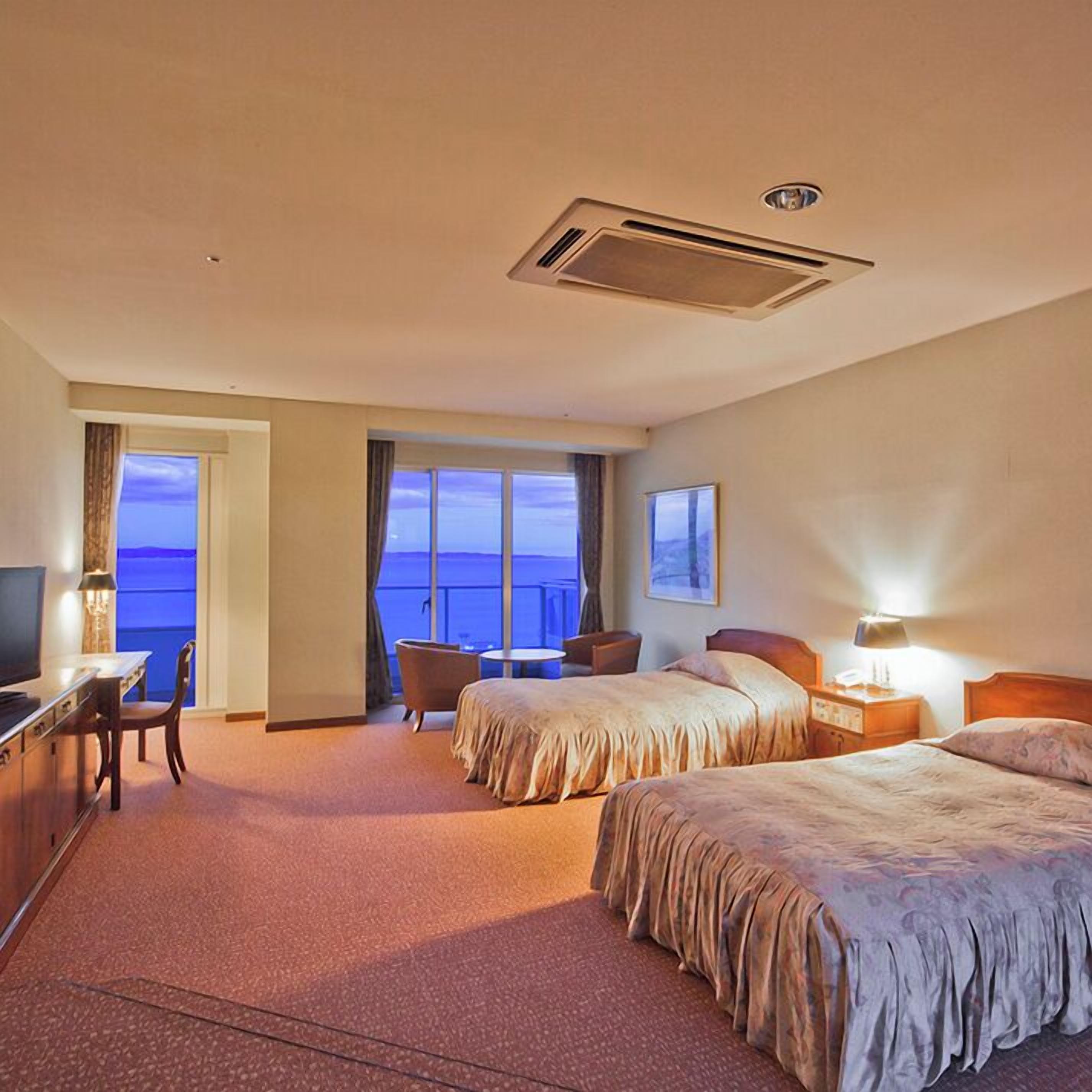 Een hotelkamer met twee bedden, een bureau met stoel, een televisie en een groot raam met uitzicht op zee.