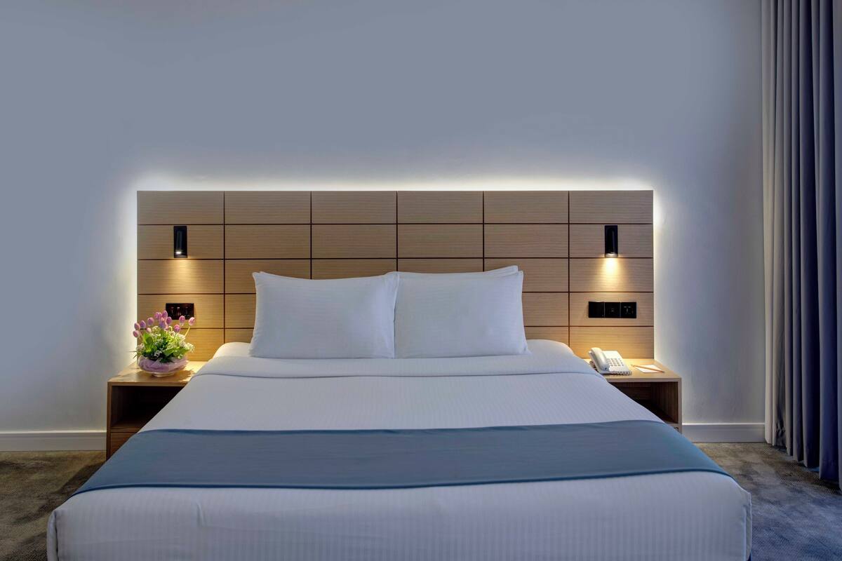 A modern hotel room with a large bed, a bedside table with a lamp, and a small table with a vase of flowers.