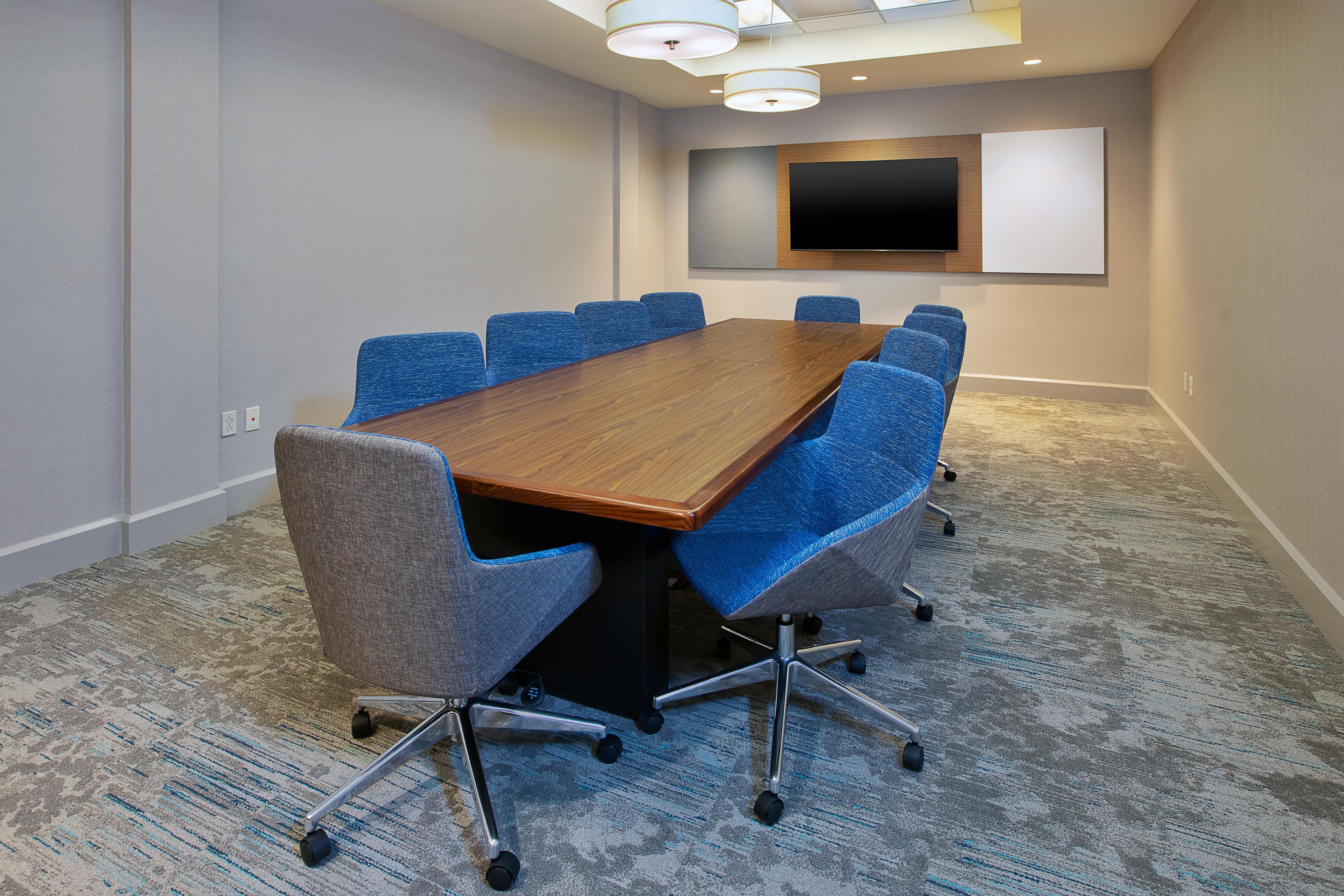 A conference room with a long wooden table, blue chairs, a flat-screen TV, and a carpeted floor.