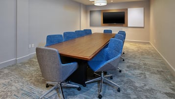 A conference room with a long wooden table, blue chairs, a flat-screen TV, and a carpeted floor.