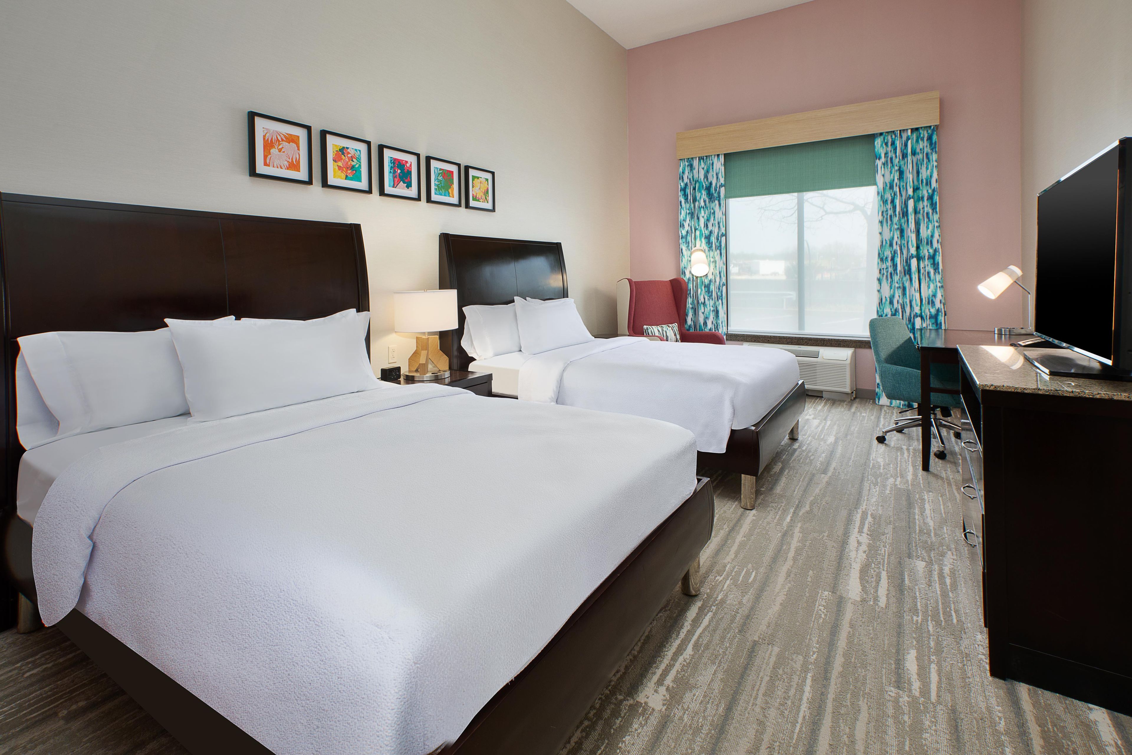 A hotel room with two beds, a desk, a TV, and a window with curtains.