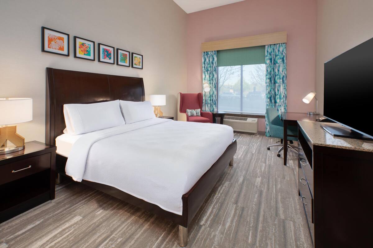 A hotel room with a bed, a desk, a chair, a TV, and artwork on the wall.
