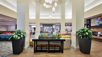 A spacious lobby with a central dining area, large potted plants, and a prominent shop sign.
