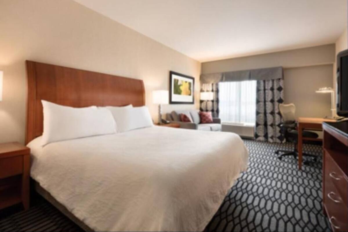 A hotel room with a large bed, a desk, a chair, a TV, and a window with curtains.