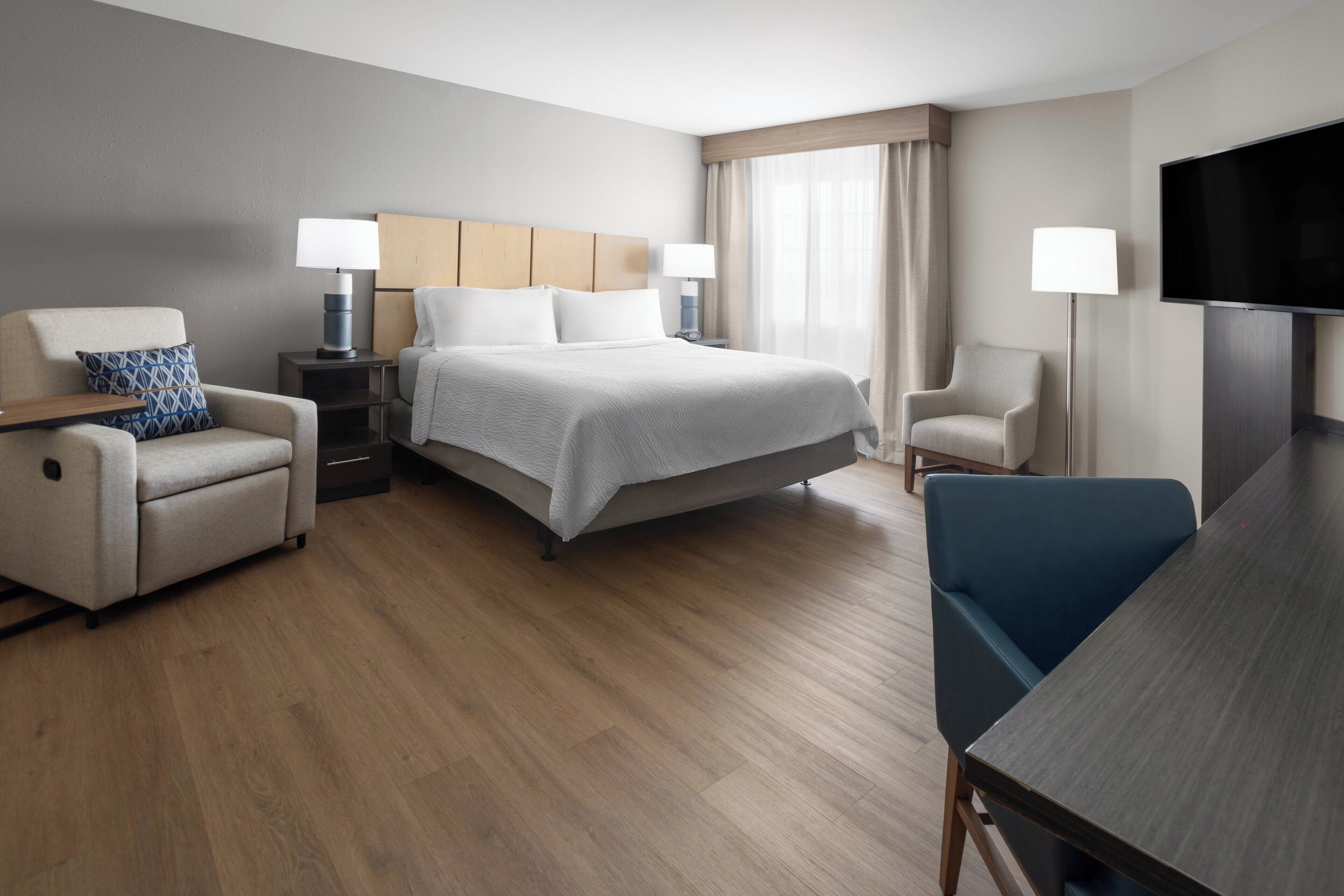 Une chambre d’hôtel comprenant un lit, deux fauteuils, un bureau, une télévision et une fenêtre avec des rideaux.
