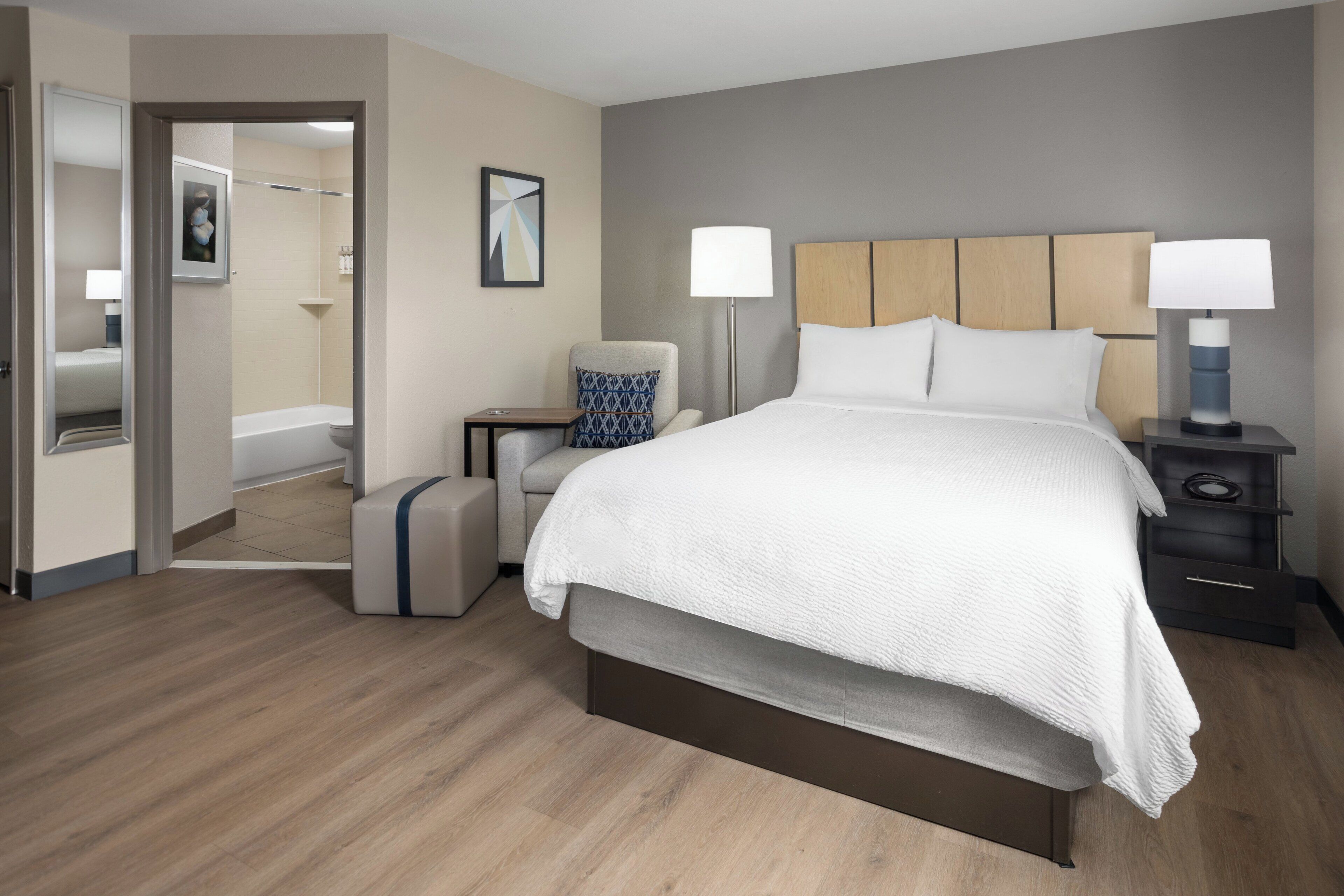 A modern hotel room with a large bed, bedside lamps, a chair, a small table, and a mirror.