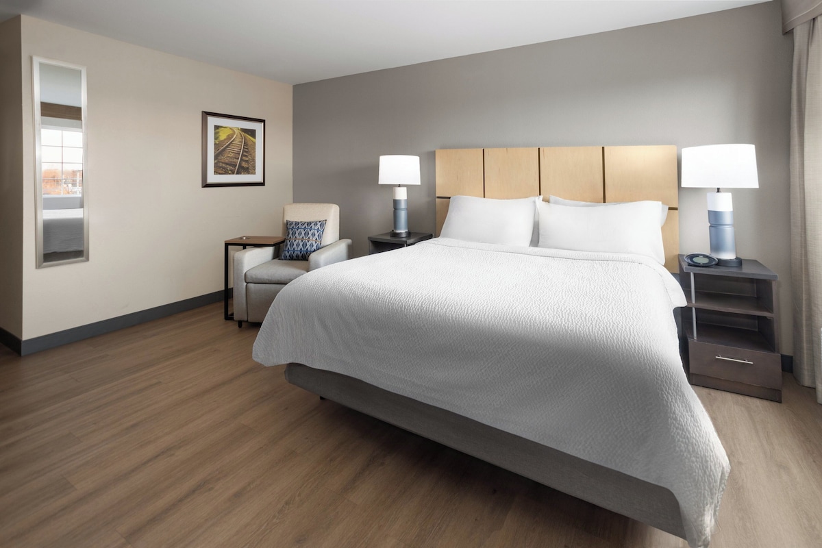 Quarto de hotel com uma cama grande, duas mesas de cabeceira com luminárias, uma cadeira, uma pequena mesa e um quadro na parede.