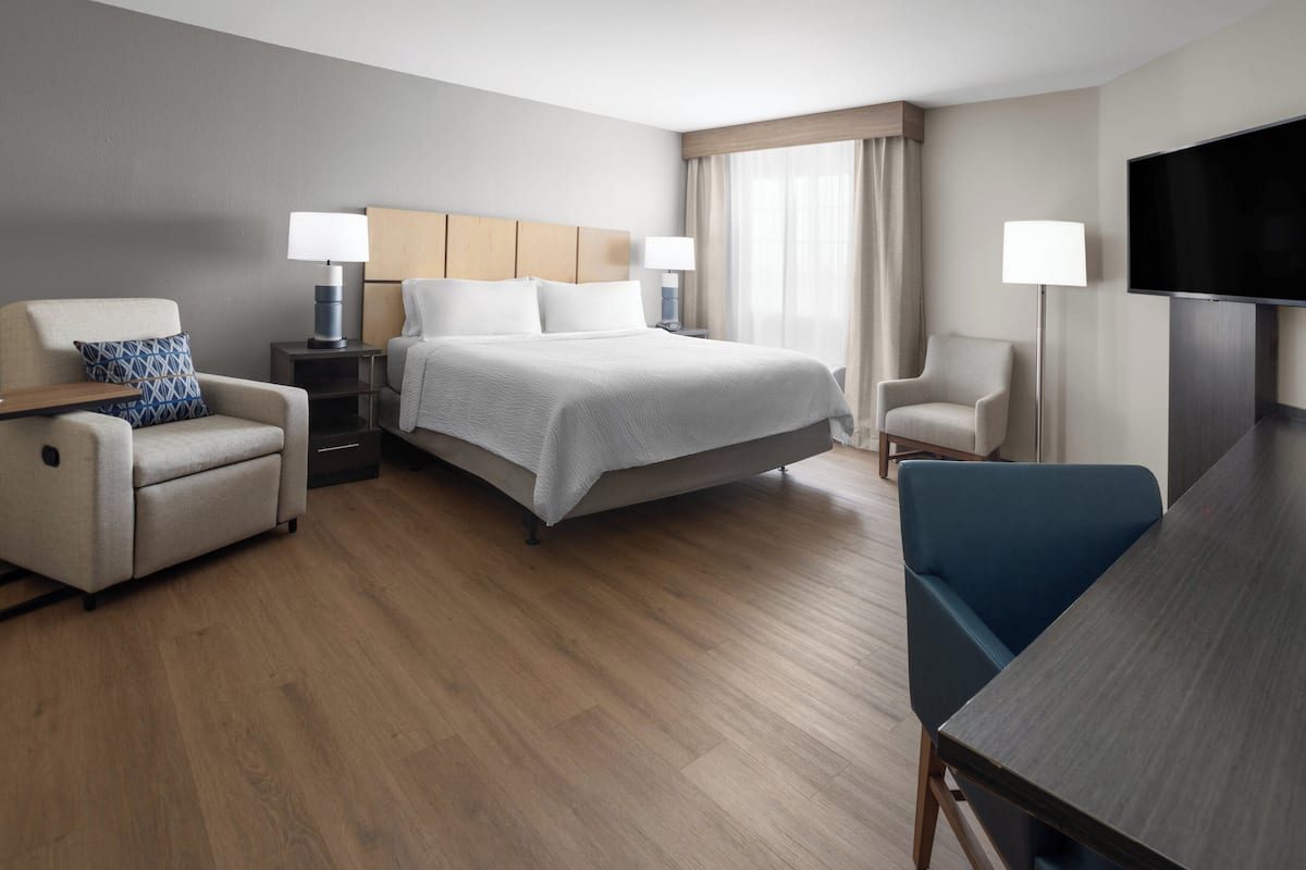 Ein Hotelzimmer mit einem Bett, zwei Sesseln, einem Schreibtisch, einem Fernseher und einem Fenster mit Vorhängen.