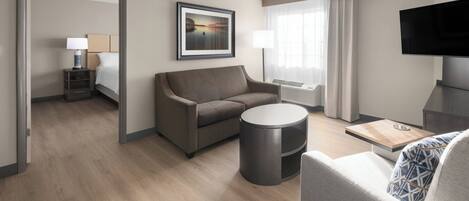 A modern hotel room with a sofa, a round coffee table, a TV, and a bed with a nightstand.