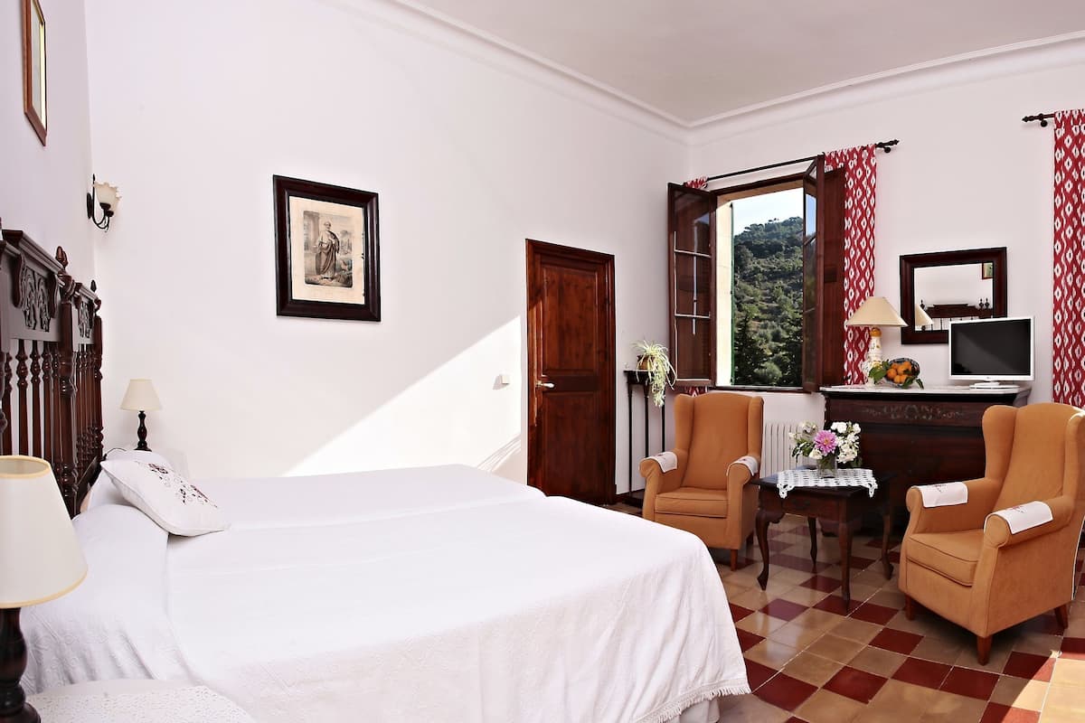 Habitación de hotel con cama, dos sillas, escritorio, televisor y ventana con vista a follaje.