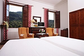 A hotel room with a bed, two armchairs, a small table with a lamp, a TV, and a view of a mountainous landscape. - Finca sa Màniga (Bunyola)