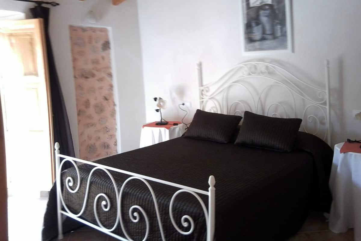 Un dormitorio con estructura de cama metálica, cabecera blanca, colcha marrón y una mesita de noche con lámpara.