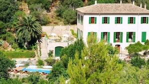 Un edificio blanco con contraventanas verdes, piscina y jardín con diversas plantas.