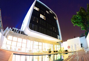 Un edificio moderno con un estanque reflectante al frente, grandes ventanales y un árbol a la derecha.