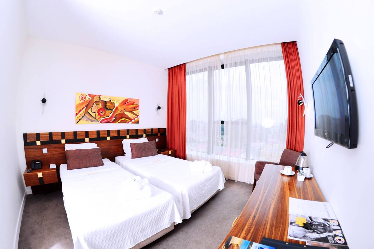 Ein Hotelzimmer mit zwei Betten, einem Holzkopfteil, einem Fernseher und einem großen Fenster mit roten Vorhängen.