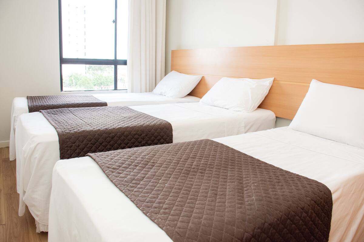 Quarto de hotel com três camas, cada uma com cabeceira de madeira e roupa de cama branca.