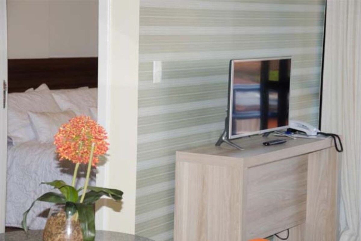 Habitación de hotel con cama, mesita de noche con televisor y un jarrón con flores.