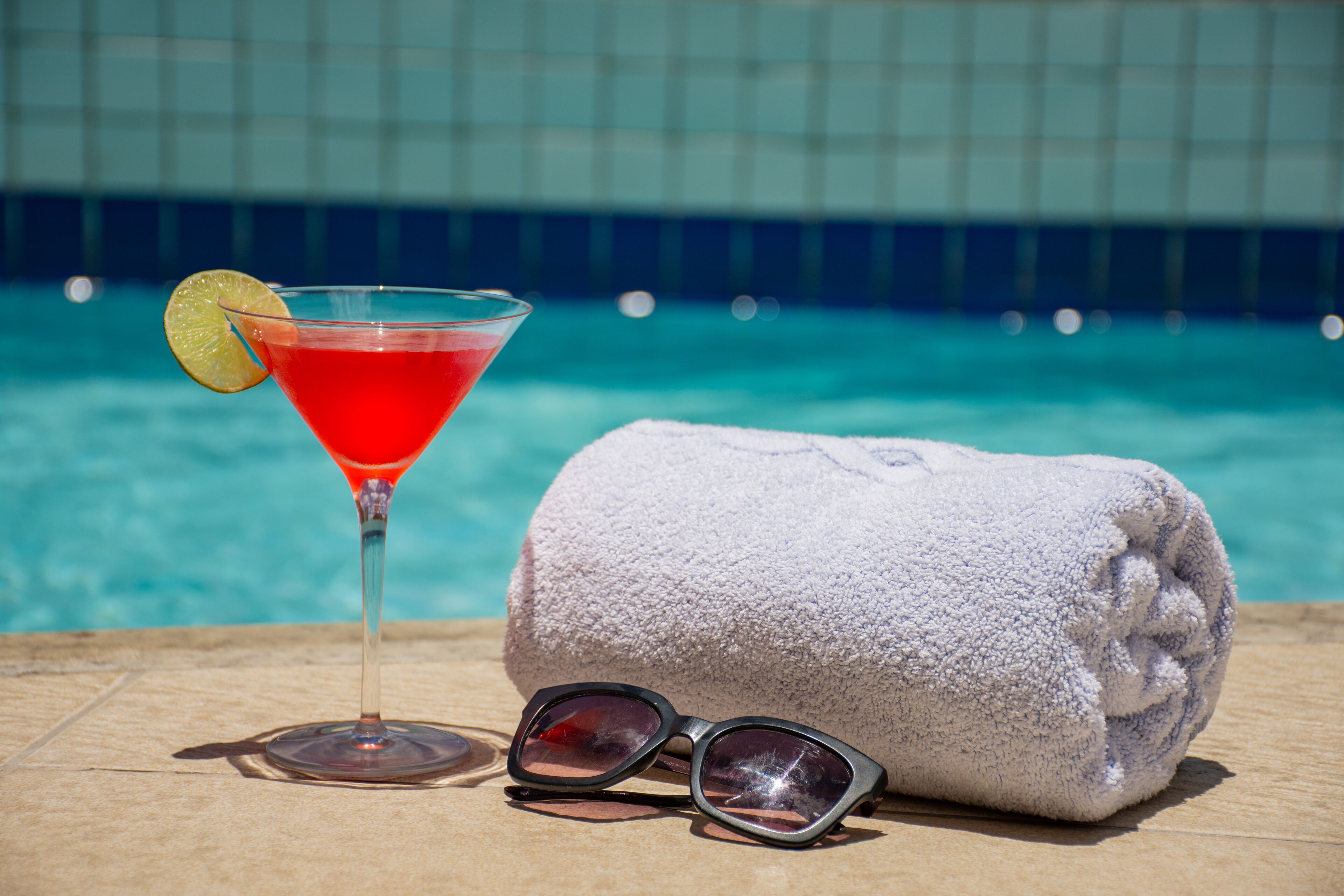 A martini glass with a red drink and a lime slice, a rolled towel, and sunglasses on a wooden surface by a pool.