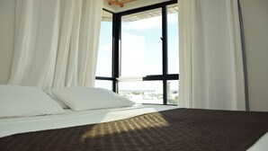 Une chambre d’hôtel avec un grand lit, des rideaux blancs et une vue sur la ville, visible à travers une grande fenêtre.