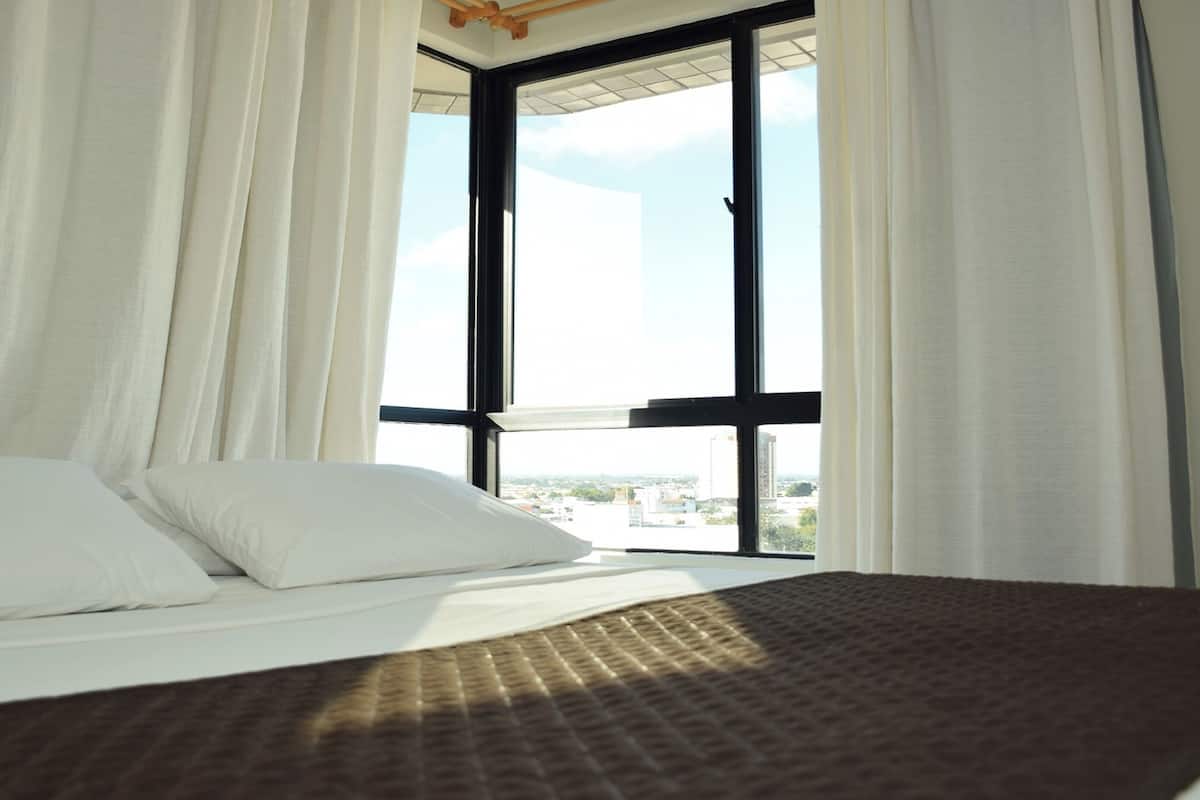 Quarto de hotel com uma cama grande, cortinas brancas e vista da cidade através de uma janela ampla.