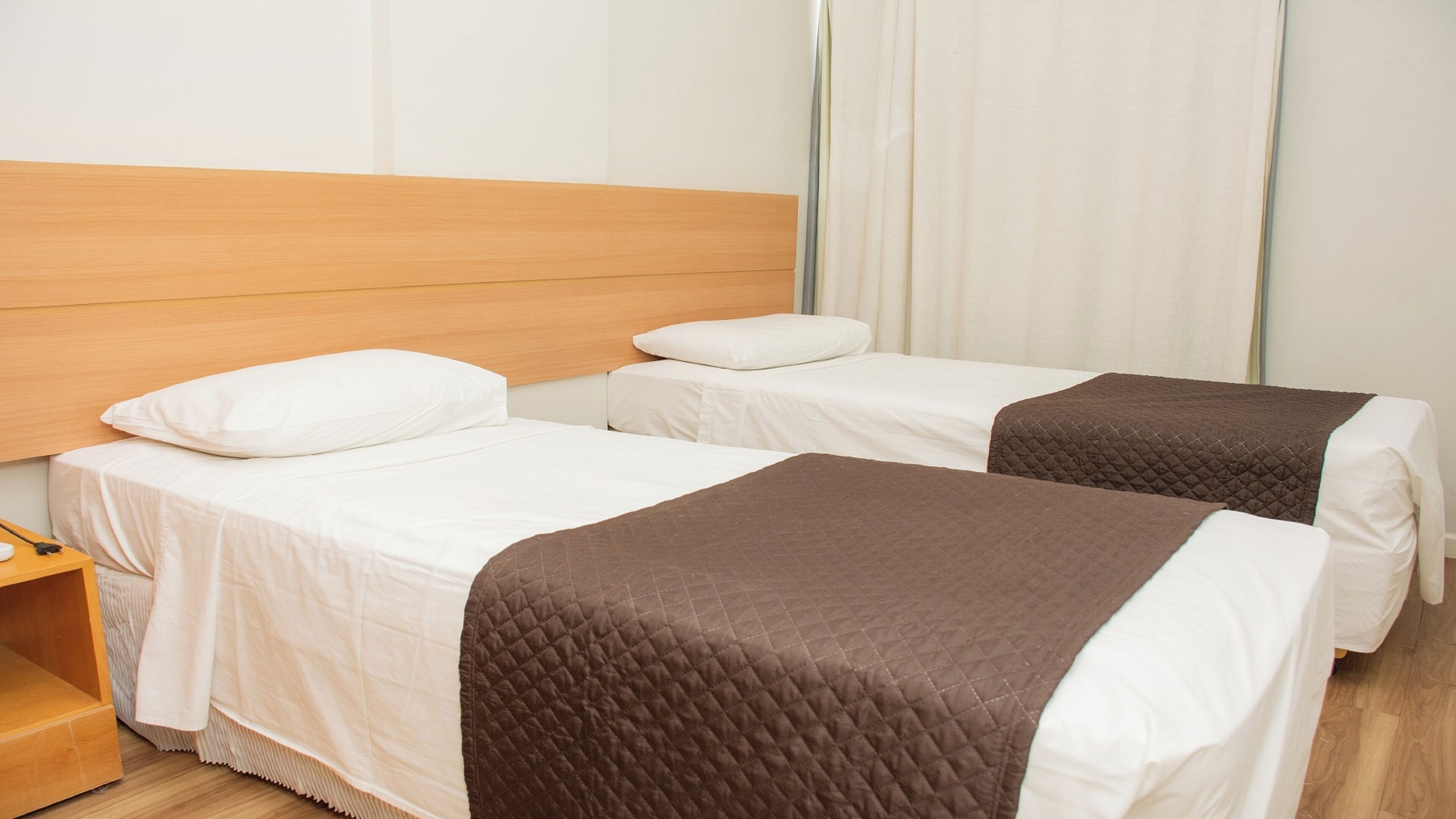 Une chambre d’hôtel avec deux lits simples, une tête de lit en bois, du linge de lit blanc et une couverture matelassée marron.