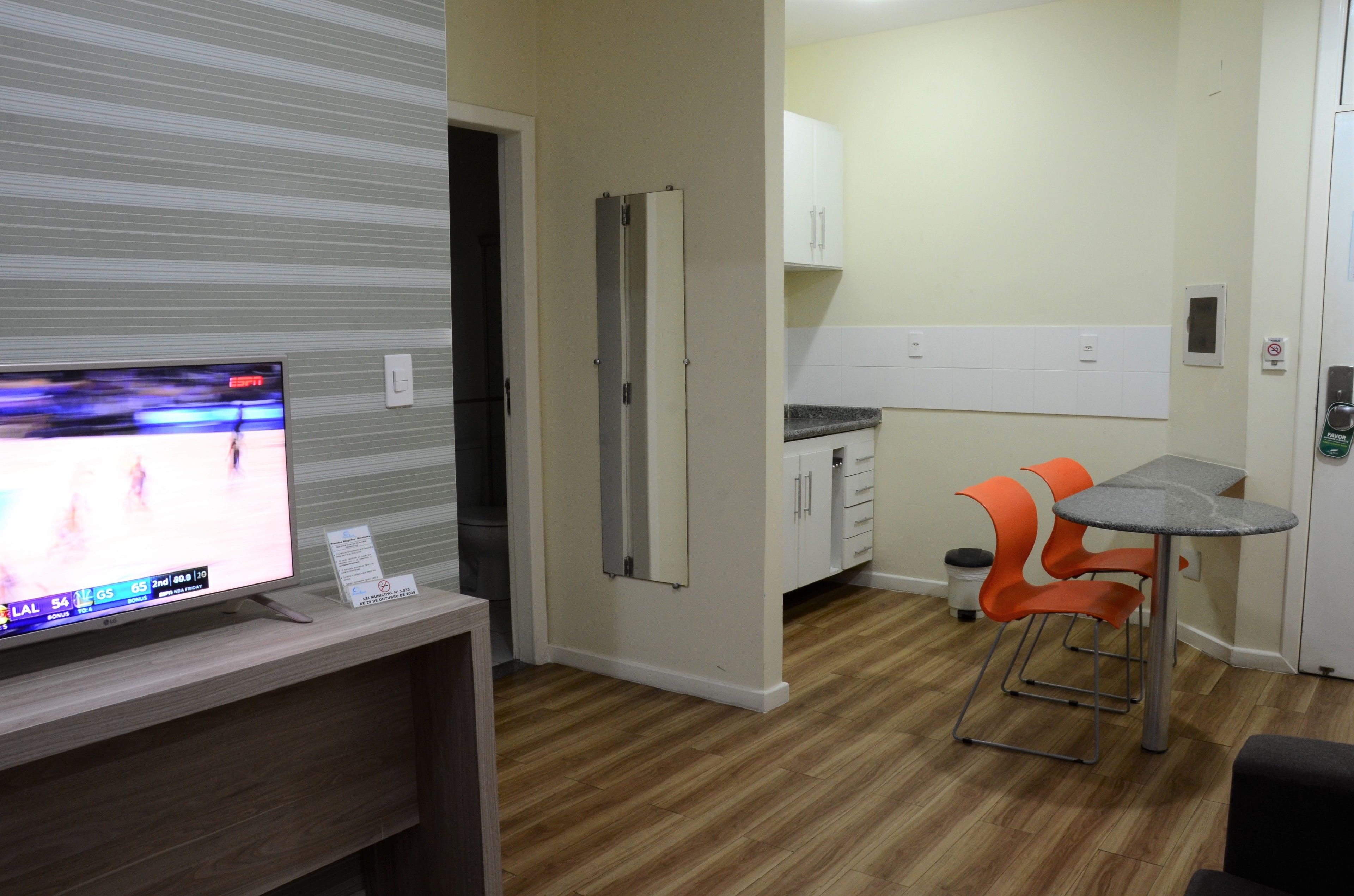 Habitación con televisor sobre un mueble de madera, cocina con armarios y encimera, y una pequeña mesa de comedor con sillas naranjas.