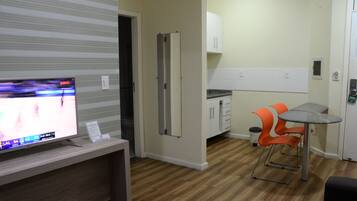 Habitación con televisor sobre un mueble de madera, cocina con armarios y encimera, y una pequeña mesa de comedor con sillas naranjas.