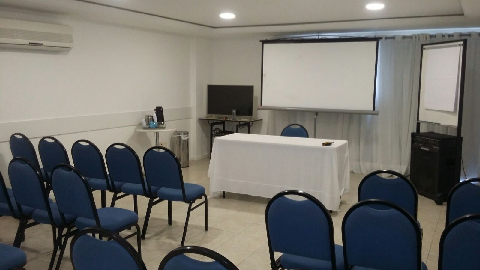 A conference room with a white table, blue chairs, a projector screen, and a television.