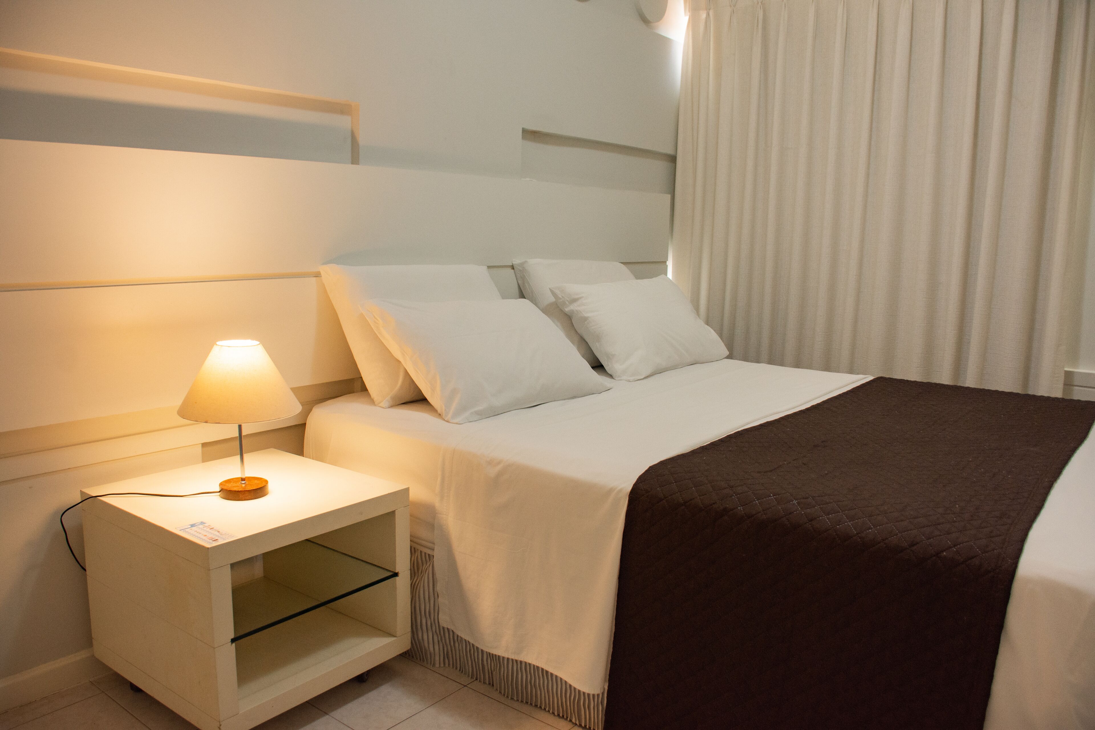 Een net opgemaakt bed met witte lakens en een bruine plaid, een nachtkastje met een lamp, en een raam met gordijnen.