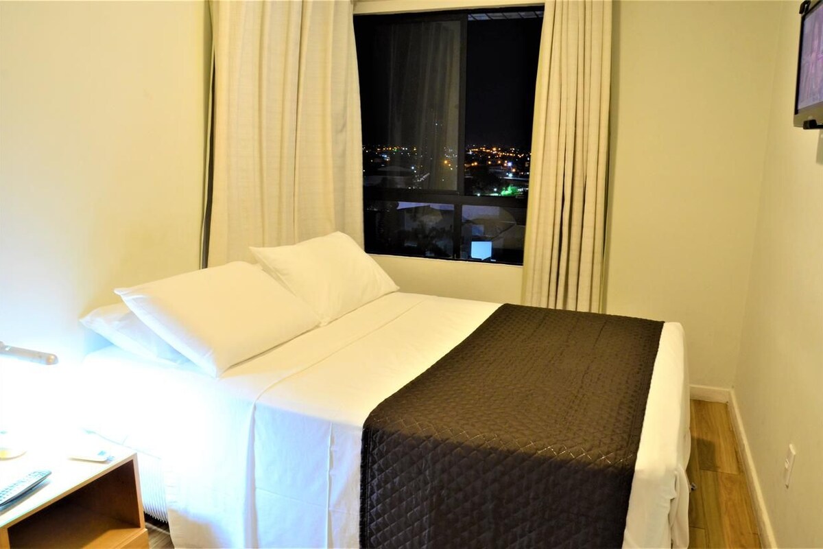 Una habitación de hotel con cama, ventana con cortinas y vista nocturna de la ciudad.