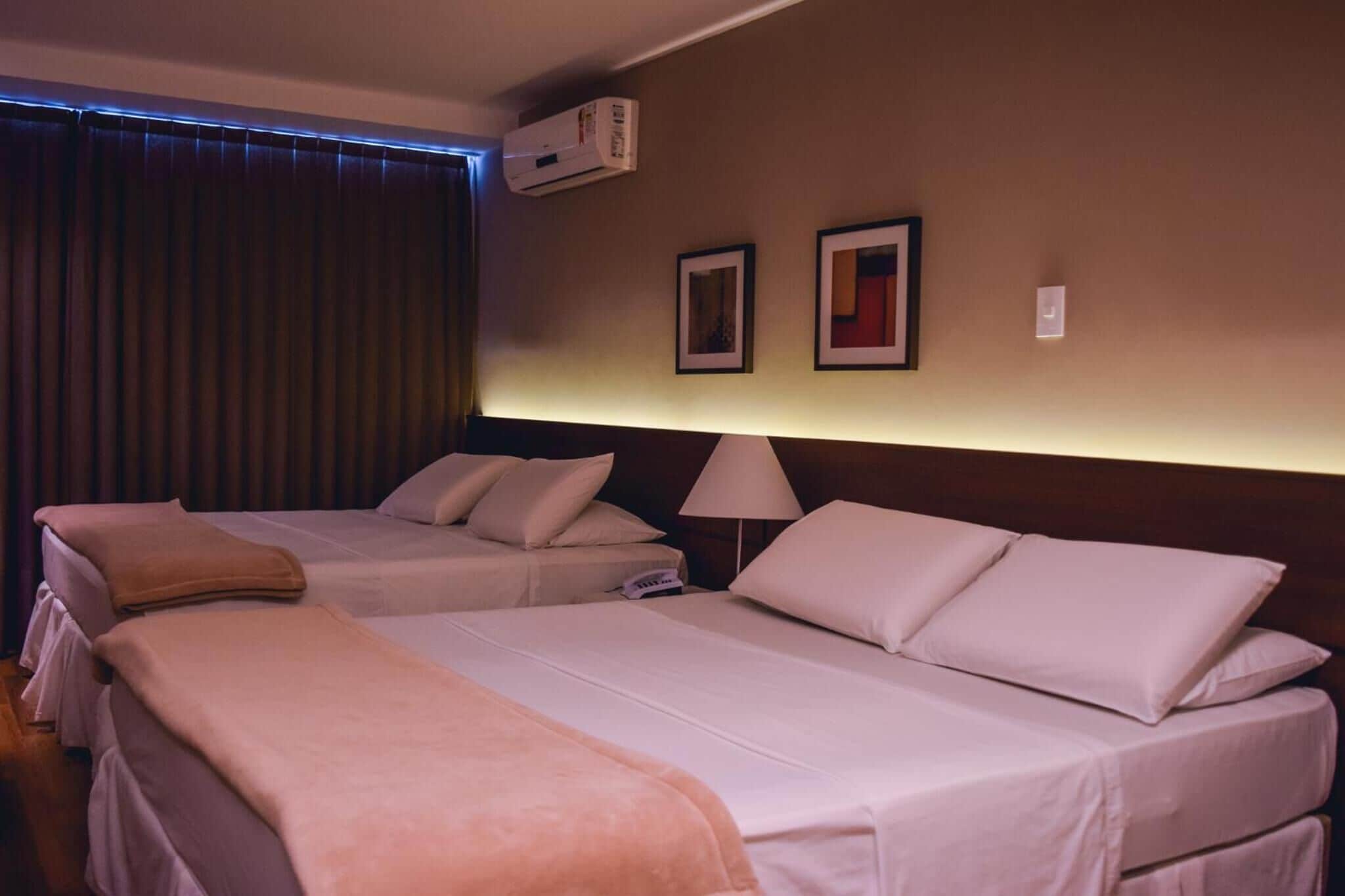 A hotel room with two beds, a bedside lamp, an air conditioning unit, and two framed pictures on the wall.