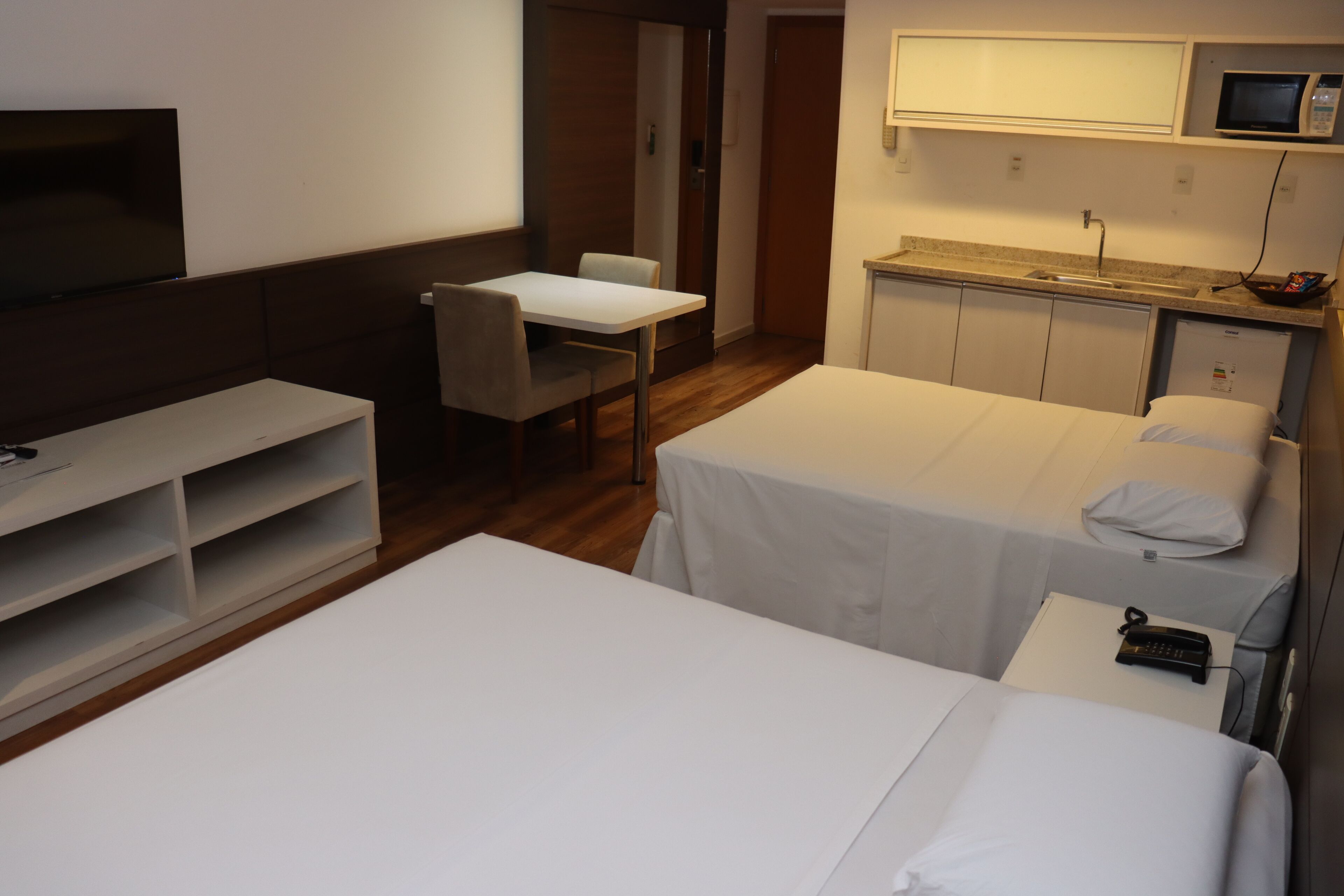 Quarto de hotel com duas camas de solteiro, uma escrivaninha, uma cadeira, televisão, micro-ondas e uma pequena cozinha equipada.
