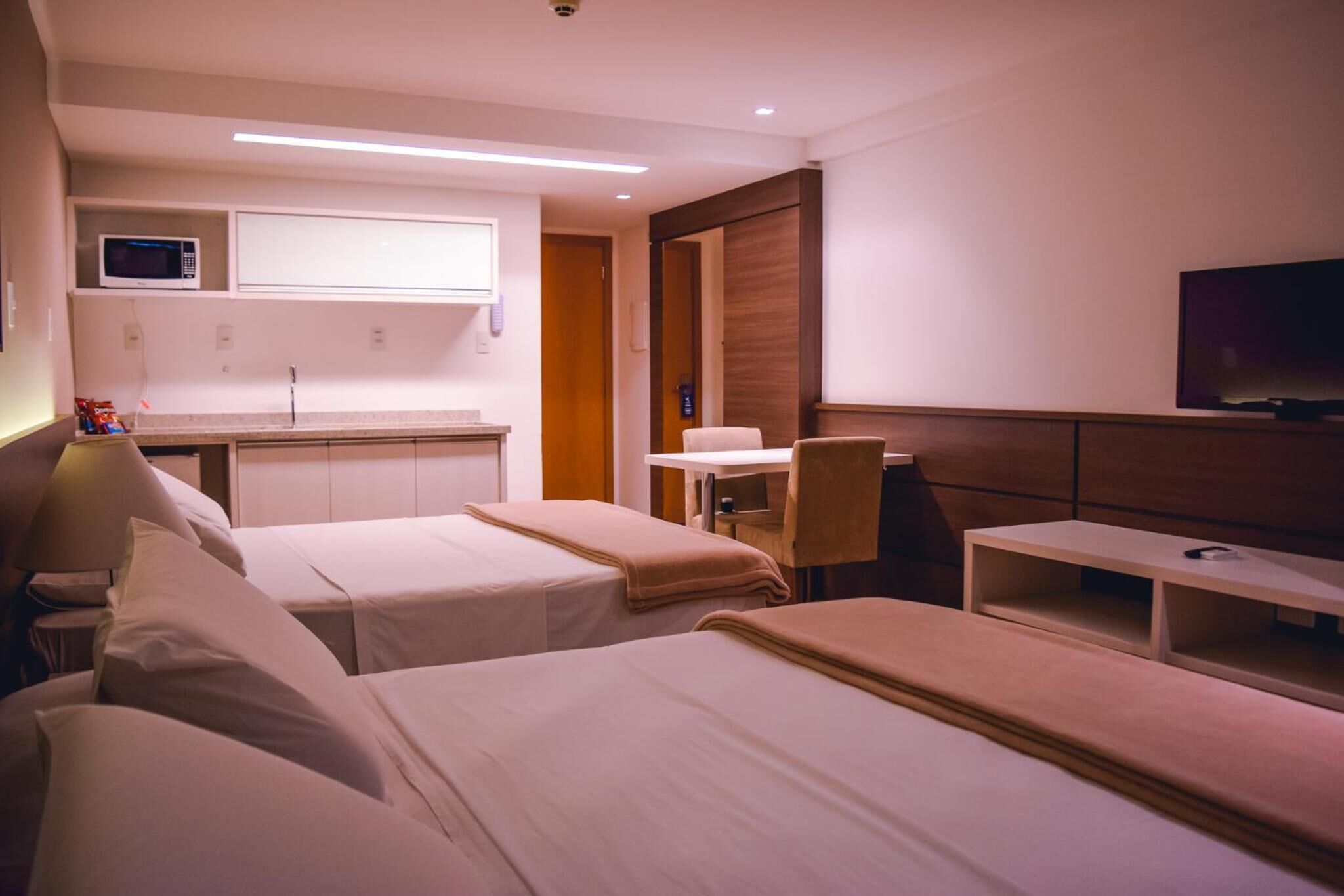 A hotel room with two beds, a desk, a TV, a microwave, and a small kitchenette.