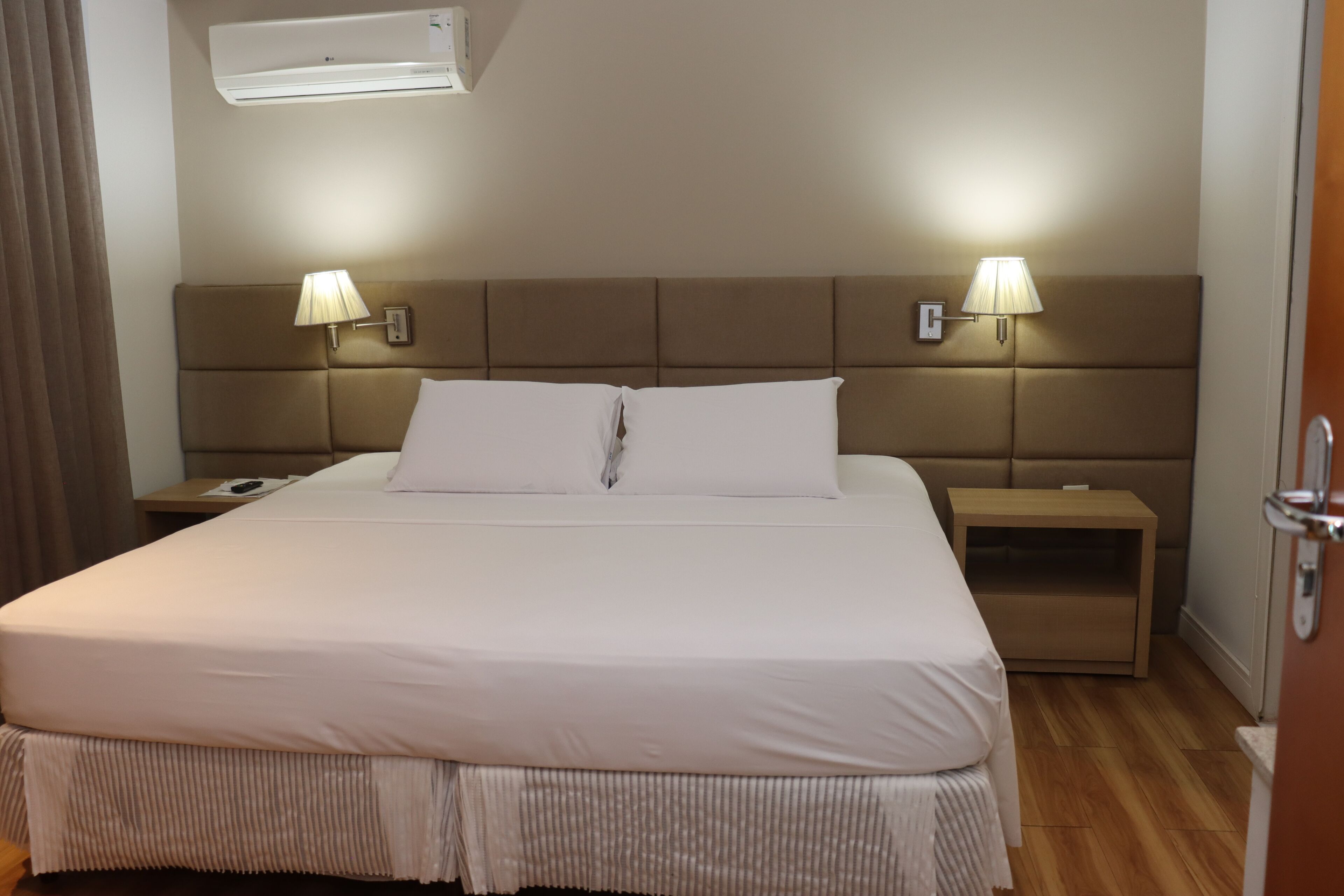 A hotel room with a large bed, two bedside lamps, a wooden nightstand, and an air conditioning unit on the wall.
