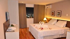 Una habitación de hotel con una cama bien hecha, una mesita de noche con lámpara, una ventana con cortinas y vistas a la ciudad.