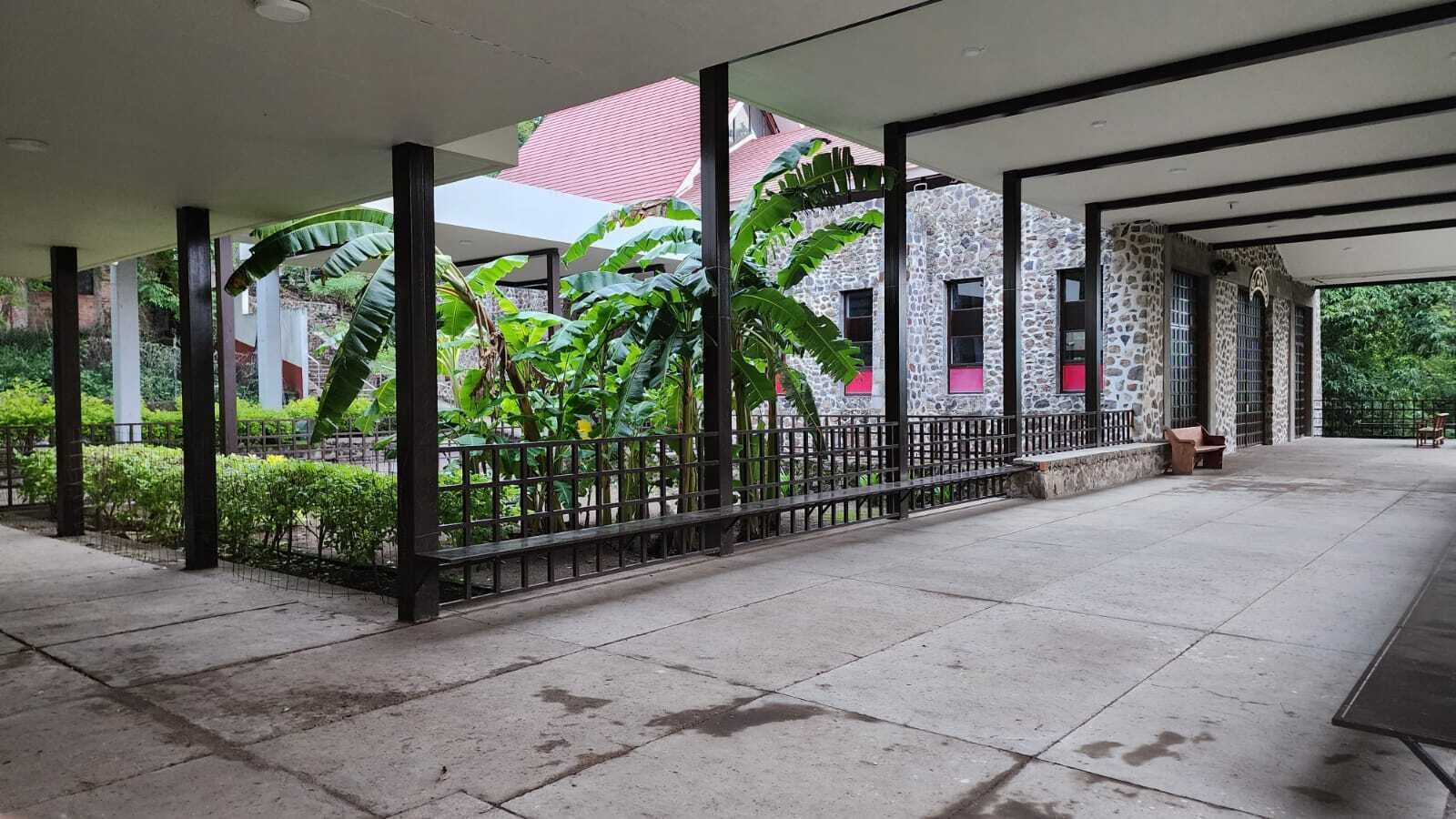 A covered outdoor area with a stone building, a metal fence, and lush greenery.