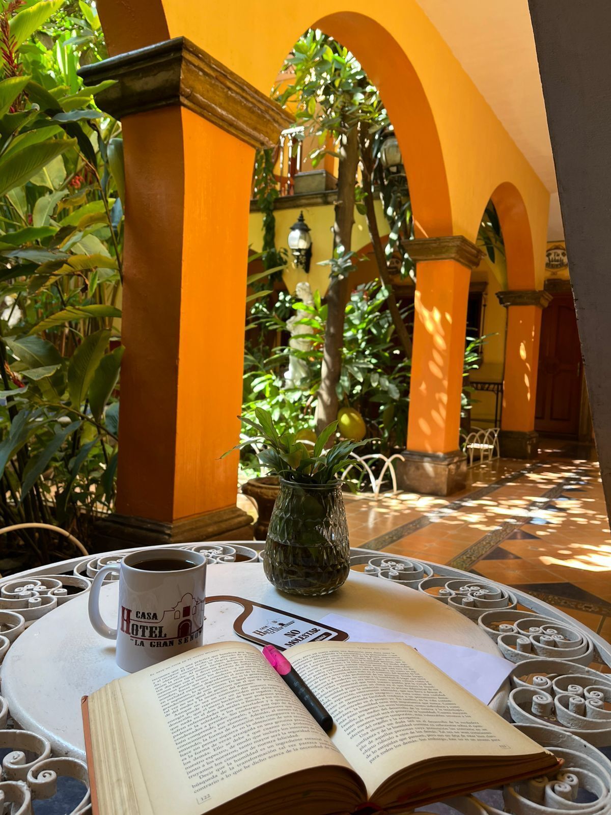 Un libro, una taza y una planta en maceta sobre una mesa en un patio soleado con arcos y follaje.