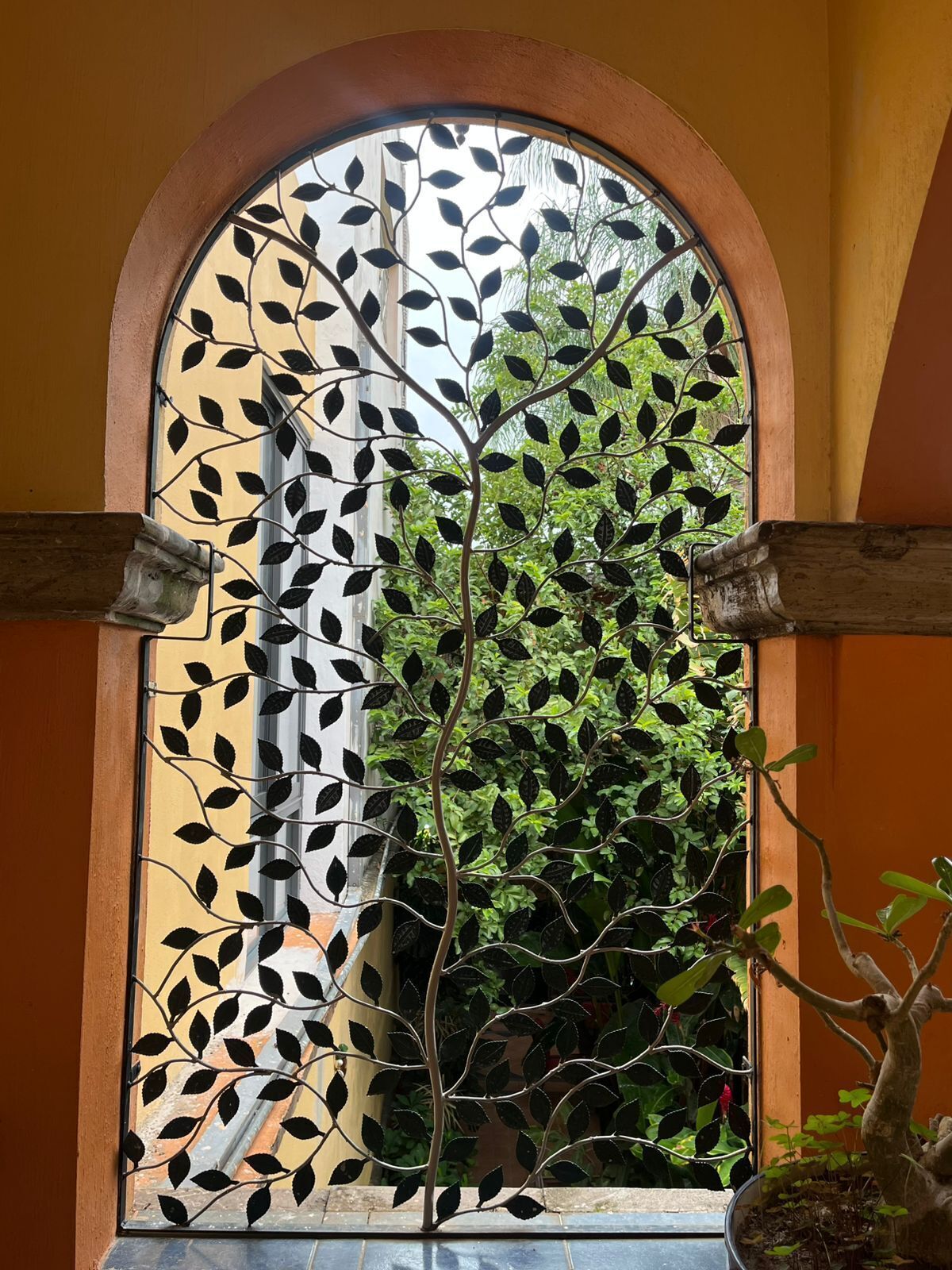 A window with a decorative metal leaf pattern, a plant in the foreground, and a view of greenery outside.