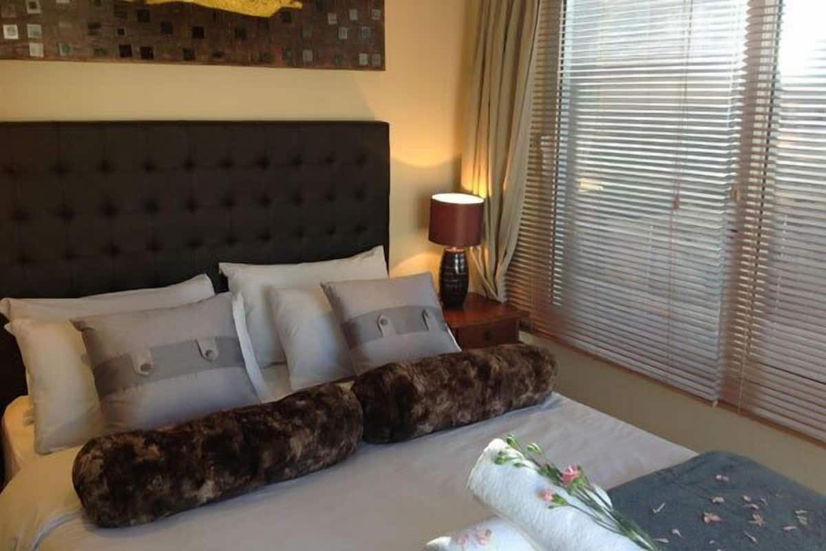 Una cama bien hecha con un cabecero oscuro, varios almohadones y un cojín decorativo. Se ve una lámpara y una ventana con persianas en el fondo.
