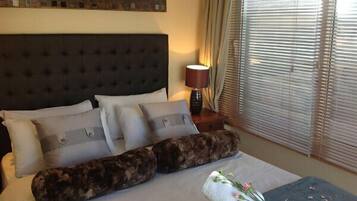 A neatly made bed with a dark headboard, multiple pillows, and a decorative pillow. A lamp and a window with blinds are visible in the background.